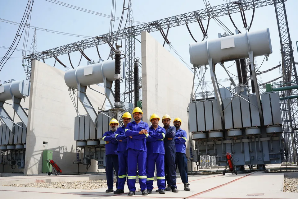 Six NationalGrid personnel in blue workwear and yellow hard hats standing in front of substation equipment